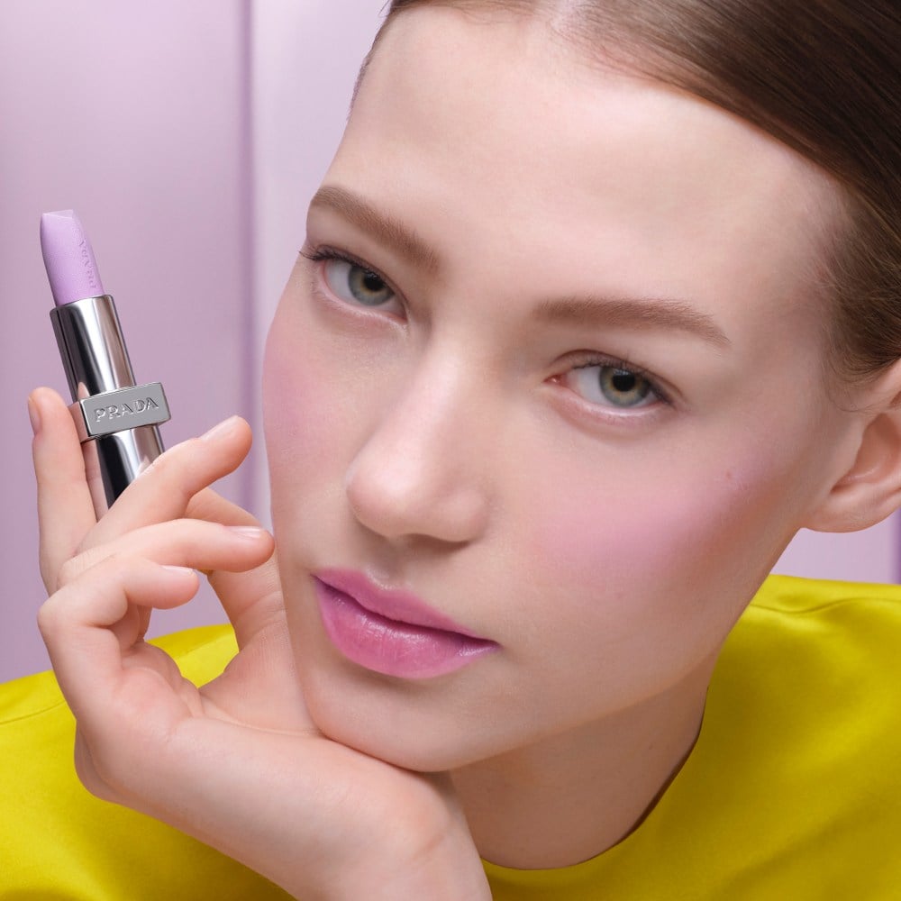 Olga (Caucasian Model) is wearing Prada Balm in shade Wisteria Gleams with product in hand on a lilac background 