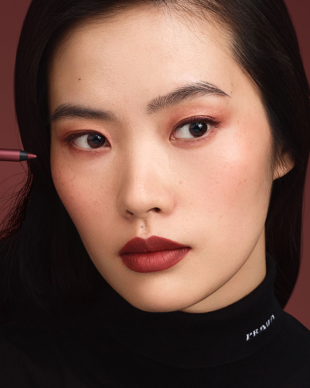 Qiangxia (Asian Skin Model) is wearing Prada Lines Eye Pencil in shade 03 Mahogany and Pradascope Mascara in shade 02 Mahogany on a burgundy background.