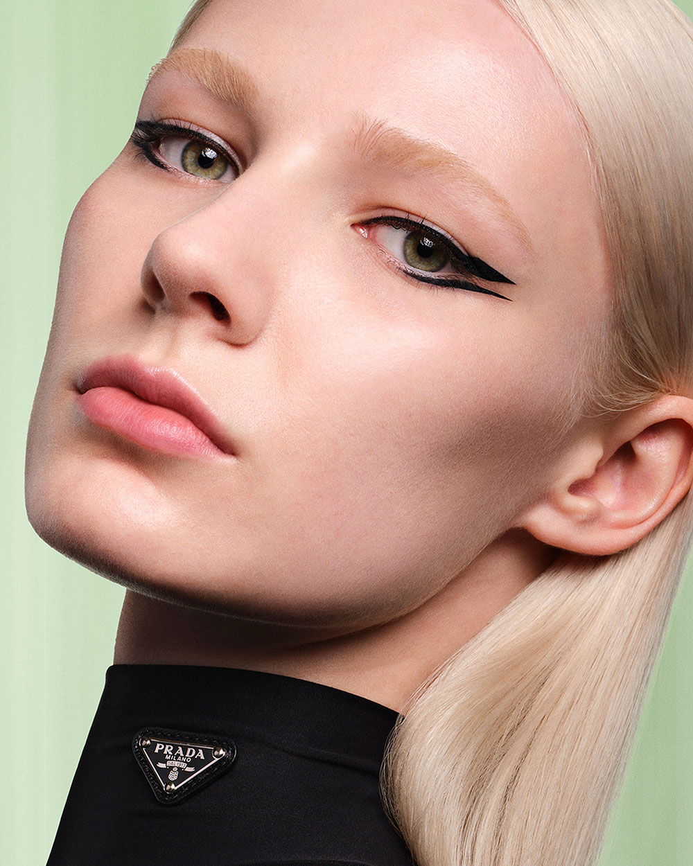 Lera (Caucasian model) is wearing Prada Lines Eye Pencil in shade 01 Nero and Pradascope Mascara in shade 01 Nero on a light green background.