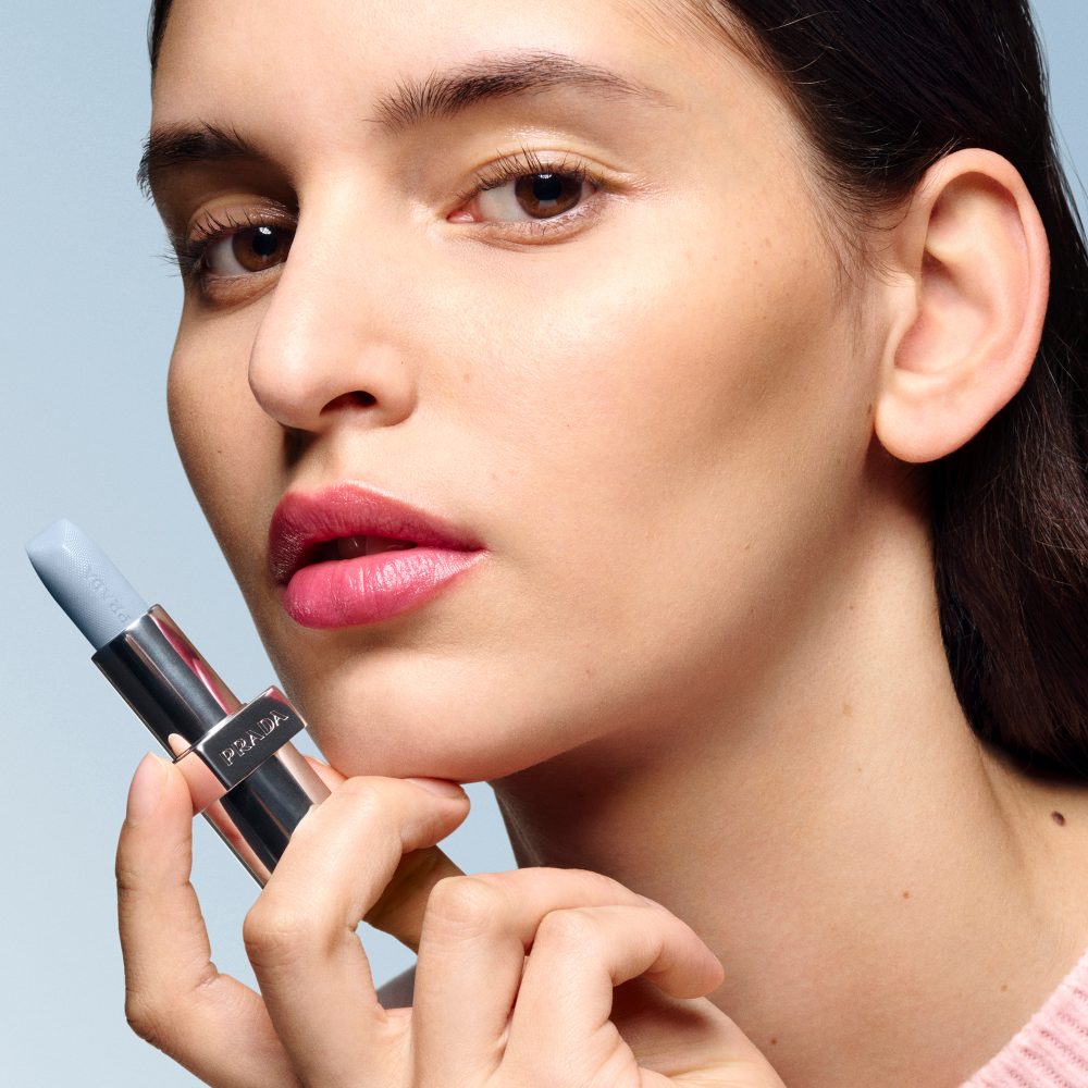 Larissa (European Model) is wearing Prada Balm in shade Astral Pink with product in hand on a light blue background 