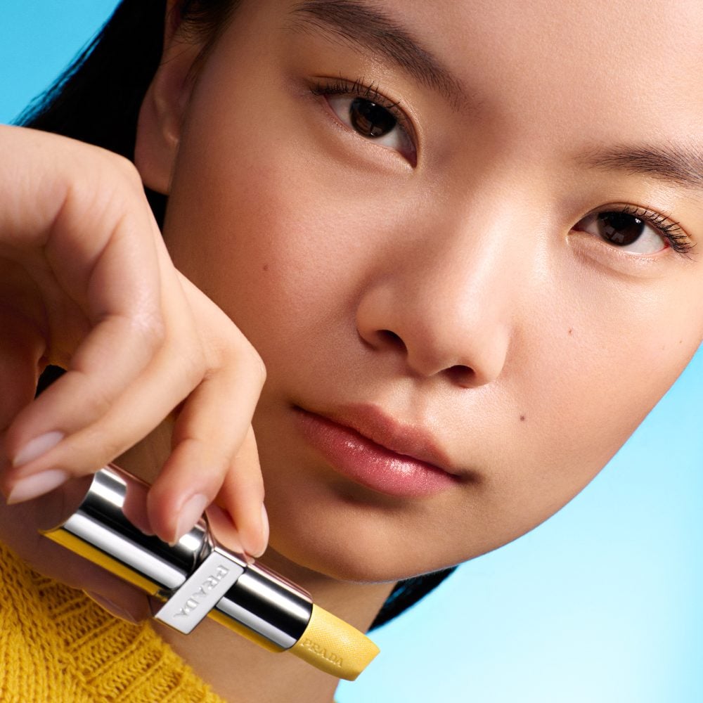 Lynn (Asian Model) is wearing Prada Balm in shade Banana Yellow with product in hand on a cyan blue background 